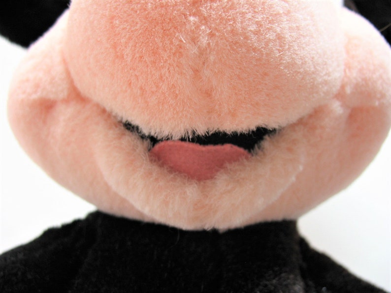 May include: Close-up of a plush toy's face. The toy has a pink nose and mouth, with a black mouth and a pink tongue.