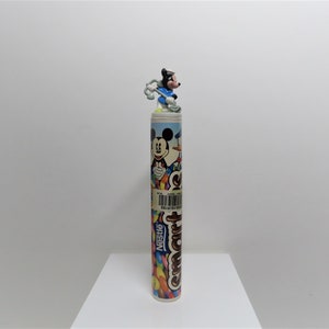 Peut inclure: Un tube coloré de bonbons Smarties Nestle avec une figurine de Mickey Mouse sur le dessus. Le tube présente une image de bande dessinée de Mickey Mouse, Donald Duck et Dingo.