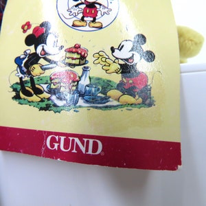 Op de afbeelding: Een geel pluche speelgoed met een rood-zwart geruit overhemd. Het speelgoed zit op een wit oppervlak. Het label op het speelgoed is rood en wit met het woord "GUND" in zwart gedrukt. Het label toont ook een cartoonbeeld van Mickey Mouse en Minnie Mouse die picknicken.