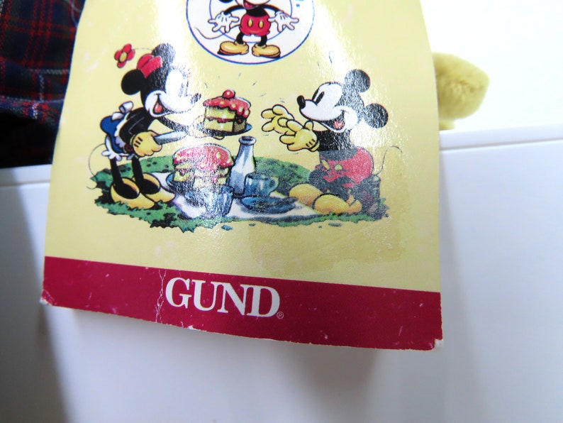 Op de afbeelding: Een geel pluche speelgoed met een rood-zwart geruit overhemd. Het speelgoed zit op een wit oppervlak. Het label op het speelgoed is rood en wit met het woord "GUND" in zwart gedrukt. Het label toont ook een cartoonbeeld van Mickey Mouse en Minnie Mouse die picknicken.