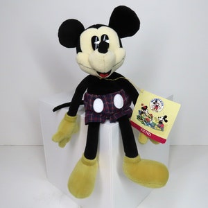 Op de afbeelding: Een zwart en geel pluche Mickey Mouse speelgoed dat een rood-blauw geruite broek draagt met witte stippen. Het speelgoed zit op een wit oppervlak en heeft een label met de tekst "Mickey Mouse" en "GUND".
