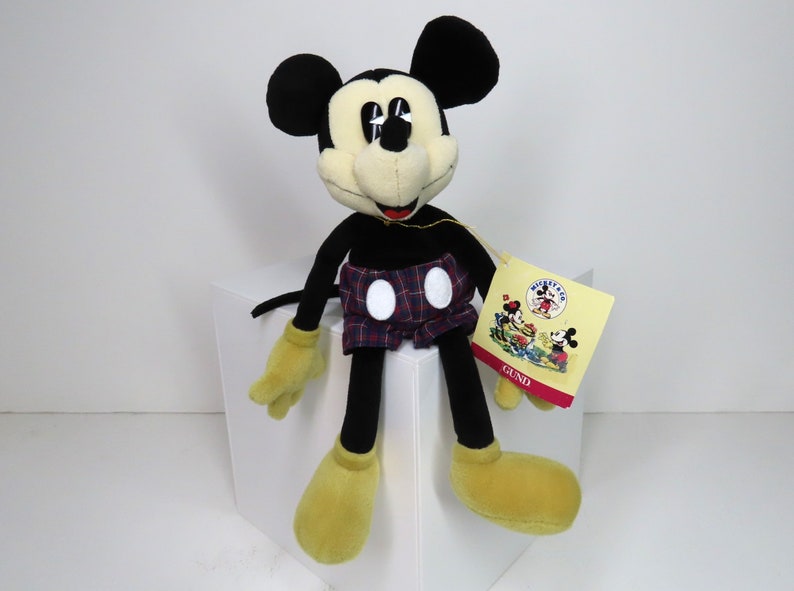 Op de afbeelding: Een zwart en geel pluche Mickey Mouse speelgoed dat een rood-blauw geruite broek draagt met witte stippen. Het speelgoed zit op een wit oppervlak en heeft een label met de tekst "Mickey Mouse" en "GUND".