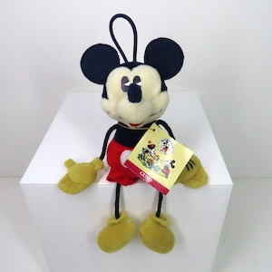 May include: A plush Mickey Mouse toy with a red and black outfit, sitting on a white surface. The toy has a tag that says "GUND" and features a vintage image of Mickey Mouse.