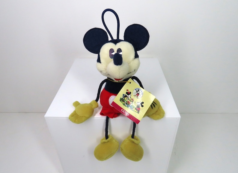May include: A plush Mickey Mouse toy with a red and black outfit, sitting on a white surface. The toy has a tag that says "GUND" and features a vintage image of Mickey Mouse.