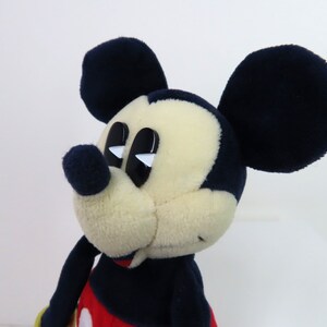 May include: A plush Mickey Mouse toy with black ears, a black nose, and large black eyes with white highlights. The toy is wearing a red shirt with a black outline and a black and white pattern.
