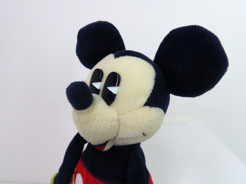 May include: A plush Mickey Mouse toy with black ears, a black nose, and large black eyes with white highlights. The toy is wearing a red shirt with a black outline and a black and white pattern.