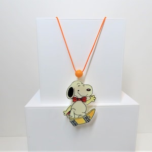 Op de afbeelding: Een witte en gele plastic ketting met een cartoon afbeelding van Snoopy op een skateboard. De ketting heeft een oranje koord en een kleine oranje kraal.