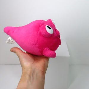 Könnte beinhalten: Rosa Plüschtier in Form eines Kleckses mit großen weißen Augen und einem kleinen Mund. Das Spielzeug wird in einer Hand gehalten.