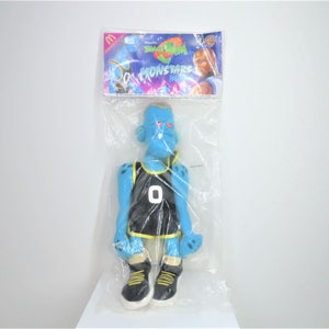 May include: A blue plush toy of a basketball player wearing a black jersey with yellow trim. The toy is wearing black trainers with yellow accents. The toy is packaged in a plastic bag with a "Space Jam" logo and a picture of a basketball player.
