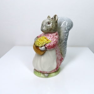 Peut inclure: Une figurine en céramique d'un écureuil gris portant une robe rose et tenant un panier de noix jaunes. L'écureuil a une queue touffue et se tient sur une base verte.