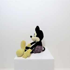 May include: A black and yellow plush Mickey Mouse toy wearing red and black plaid shorts. The toy is sitting on a white surface.