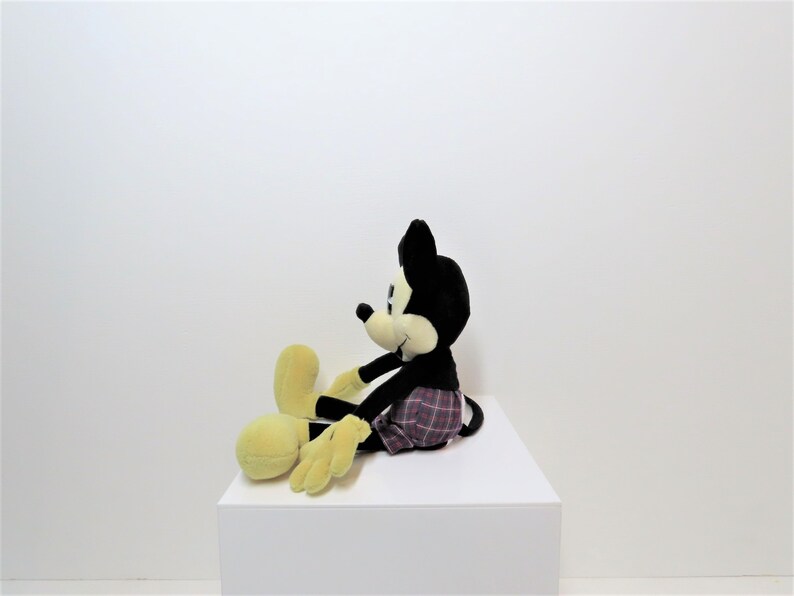 May include: A black and yellow plush Mickey Mouse toy wearing red and black plaid shorts. The toy is sitting on a white surface.