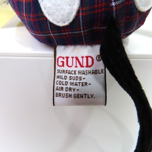 Op de afbeelding: Een rood, zwart en wit geruit knuffeldier met een zwarte staart en een wit label met de tekst "GUND&reg; SURFACE WASHABLE MILD SUDS - COLD WATER - AIR DRY - BRUSH GENTLY."