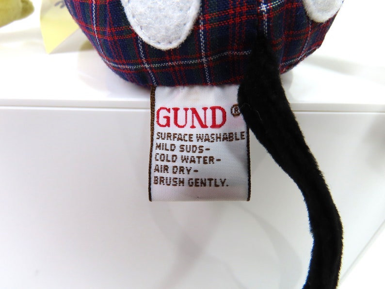 Op de afbeelding: Een rood, zwart en wit geruit knuffeldier met een zwarte staart en een wit label met de tekst "GUND&reg; SURFACE WASHABLE MILD SUDS - COLD WATER - AIR DRY - BRUSH GENTLY."
