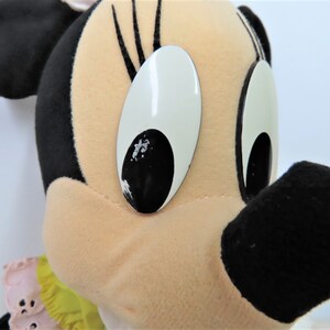 Puede incluir: Un juguete de peluche de Minnie Mouse con orejas negras, una nariz negra y grandes ojos blancos con pupilas negras. Lleva un vestido amarillo y rosa con un cuello de volantes.