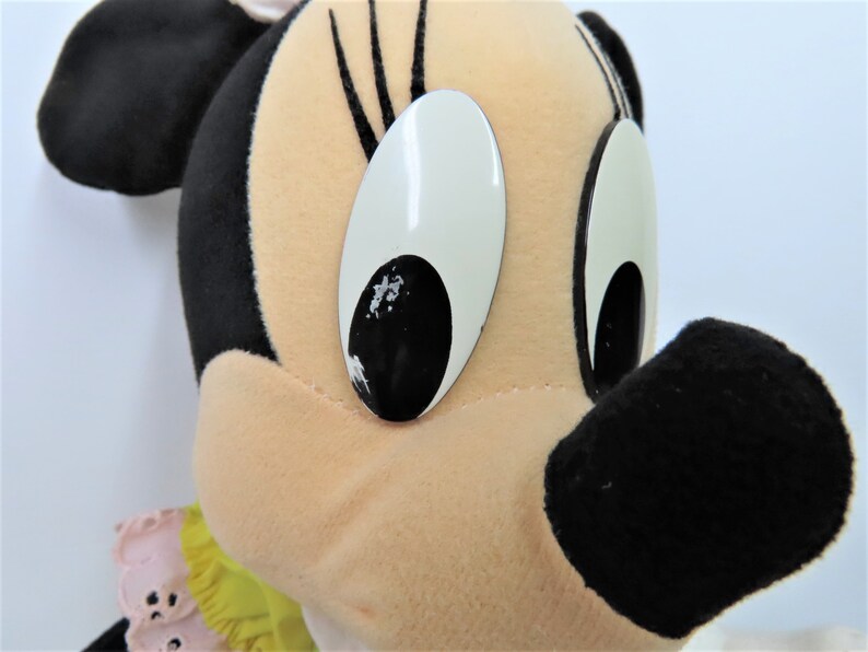 Puede incluir: Un juguete de peluche de Minnie Mouse con orejas negras, una nariz negra y grandes ojos blancos con pupilas negras. Lleva un vestido amarillo y rosa con un cuello de volantes.