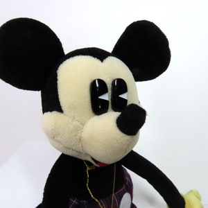Op de afbeelding: Een pluche Mickey Mouse speelgoed met zwarte oren, een zwarte neus en grote zwarte ogen met witte pupillen. Het speelgoed draagt een rood-zwart geruit overhemd en een gouden ketting.