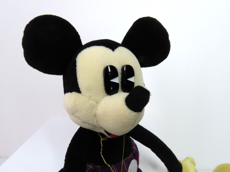 Op de afbeelding: Een pluche Mickey Mouse speelgoed met zwarte oren, een zwarte neus en grote zwarte ogen met witte pupillen. Het speelgoed draagt een rood-zwart geruit overhemd en een gouden ketting.