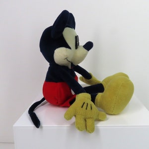 May include: A plush Mickey Mouse toy sitting with his legs crossed. He is wearing red shorts, black shoes, and yellow gloves. His ears are black and his face is white.