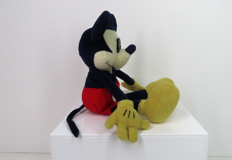 May include: A plush Mickey Mouse toy sitting with his legs crossed. He is wearing red shorts, black shoes, and yellow gloves. His ears are black and his face is white.