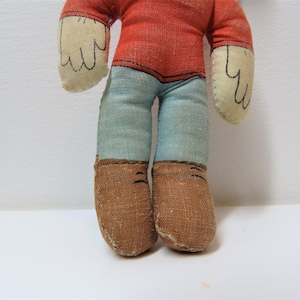 K&ouml;nnte beinhalten: Eine Vintage-Stoffpuppe mit rotem Shirt, blauer Hose und braunen Schuhen. Die Puppe hat ein einfaches, handgen&auml;htes Design.