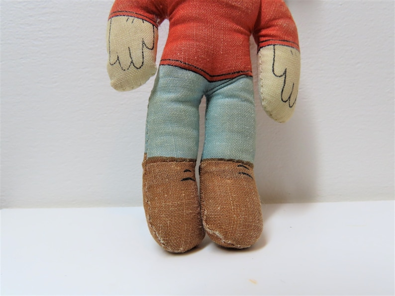 K&ouml;nnte beinhalten: Eine Vintage-Stoffpuppe mit rotem Shirt, blauer Hose und braunen Schuhen. Die Puppe hat ein einfaches, handgen&auml;htes Design.