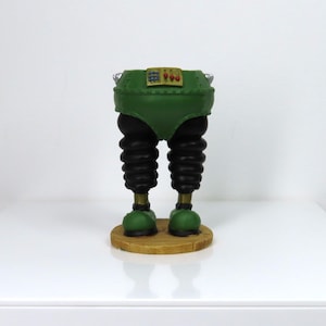 May include: A quirky figurine depicting a lower body with a green torso, black, concertina-style legs, and green feet. It rests on a circular wooden base. The figurine has a gold-coloured belt. The overall design is unusual and eye-catching.