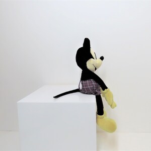 May include: A black and yellow plush Mickey Mouse toy wearing a red and black plaid shorts. The toy is sitting on a white surface.