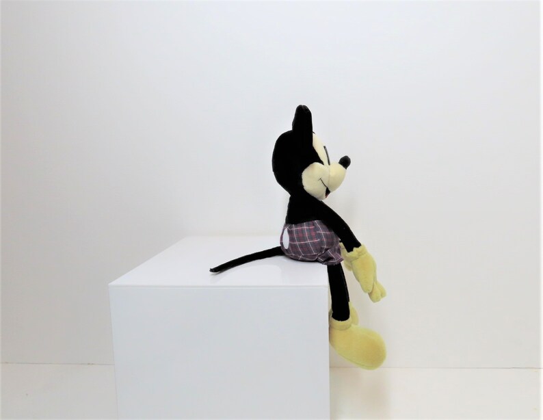 May include: A black and yellow plush Mickey Mouse toy wearing a red and black plaid shorts. The toy is sitting on a white surface.