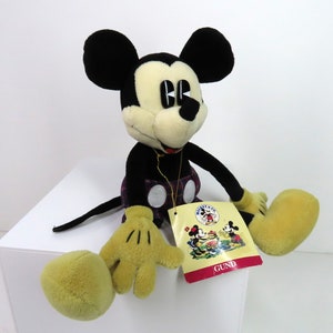 Op de afbeelding: Een pluche Mickey Mouse speelgoed dat een geruite kilt draagt en zit met zijn benen uitgestrekt. Het speelgoed heeft een zwart-gele kleurstelling en houdt een label vast met de tekst "GUND".
