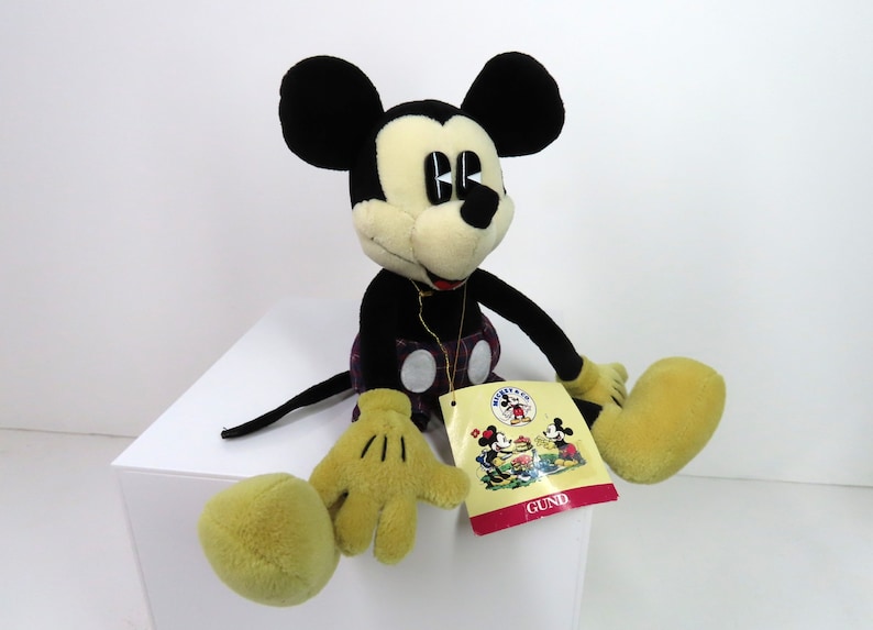 Op de afbeelding: Een pluche Mickey Mouse speelgoed dat een geruite kilt draagt en zit met zijn benen uitgestrekt. Het speelgoed heeft een zwart-gele kleurstelling en houdt een label vast met de tekst "GUND".