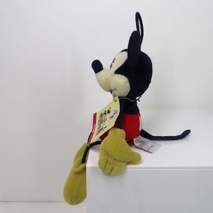 May include: A plush Mickey Mouse toy sitting on a white surface. The toy is wearing red shorts, yellow gloves, and black shoes. The toy has a blue body and a black nose. The toy has a tag attached to its right leg.