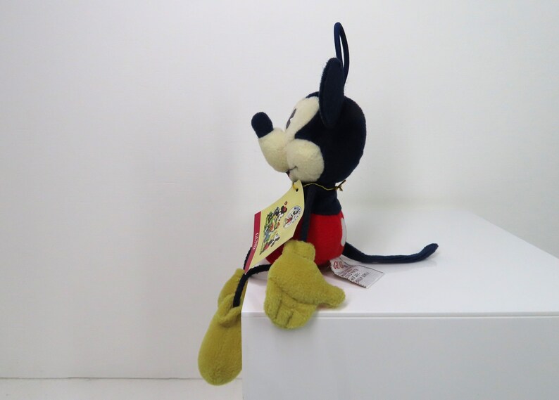 May include: A plush Mickey Mouse toy sitting on a white surface. The toy is wearing red shorts, yellow gloves, and black shoes. The toy has a blue body and a black nose. The toy has a tag attached to its right leg.