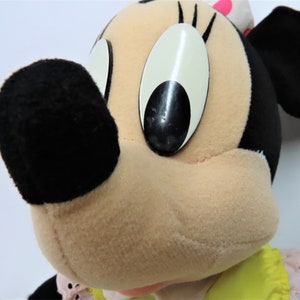 Puede incluir: Un juguete de peluche de Minnie Mouse, la novia de Mickey Mouse. Lleva un vestido amarillo con ribetes rosas y tiene una nariz negra y ojos grandes negros.