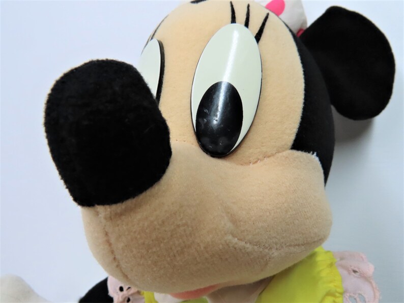 Puede incluir: Un juguete de peluche de Minnie Mouse, la novia de Mickey Mouse. Lleva un vestido amarillo con ribetes rosas y tiene una nariz negra y ojos grandes negros.