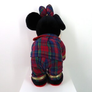 May include: A plush toy of Mickey Mouse wearing red and blue plaid pajamas with gold trim. The toy is standing on a white surface.