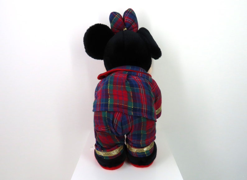 May include: A plush toy of Mickey Mouse wearing red and blue plaid pajamas with gold trim. The toy is standing on a white surface.
