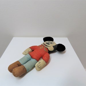 K&ouml;nnte beinhalten: Eine vintage Pl&uuml;sch-Mickey-Mouse-Puppe mit schwarzen Ohren, einem roten Shirt, einer blauen Hose und braunen Schuhen. Die Puppe liegt auf einer wei&szlig;en Oberfl&auml;che.