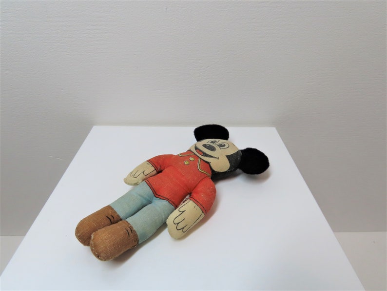 K&ouml;nnte beinhalten: Eine vintage Pl&uuml;sch-Mickey-Mouse-Puppe mit schwarzen Ohren, einem roten Shirt, einer blauen Hose und braunen Schuhen. Die Puppe liegt auf einer wei&szlig;en Oberfl&auml;che.