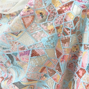 May include: Close-up of a sheer fabric with a patchwork design. The fabric features a light blue background with geometric shapes in various colors, including pink, yellow, and brown. The shapes are embellished with sequins.