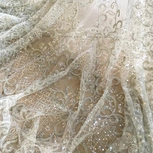May include: White beaded lace fabric with a delicate floral pattern. The fabric is sheer and has a subtle sparkle.