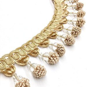 Peut inclure: Garniture décorative dorée avec un motif bouclé et des glands pendants. La garniture présente un motif tissé avec de petits glands beiges en forme de fleur et des perles claires à facettes. Idéal pour les projets de couture.