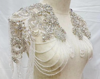 May include: A silver and crystal bridal shoulder piece with a beaded chain design. The piece is made of rhinestones and beads and is designed to be worn over a wedding dress.