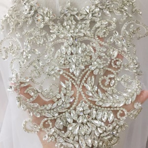 May include: A delicate, intricate design featuring a floral motif with clear rhinestones and crystals. The design is sewn onto a sheer, white mesh fabric.