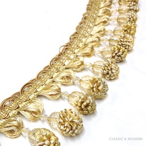 Peut inclure: Garniture dorée avec frange à perles. La frange présente de petites perles rondes transparentes et des glands dorés. La garniture est idéale pour ajouter une touche d'élégance aux rideaux, aux coussins ou à d'autres projets de décoration intérieure.