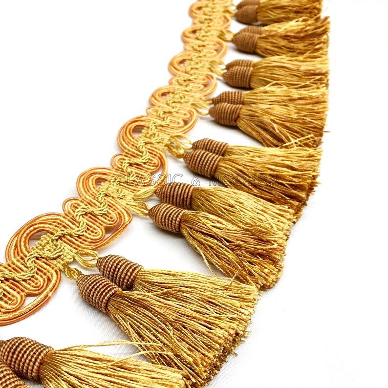 May include: Gold and brown tassel trim with a decorative woven design. The trim features a series of tassels with a looped design.