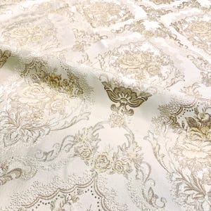 Può includere: Tessuto bianco con un motivo damascato beige con disegni floreali e a volute.
