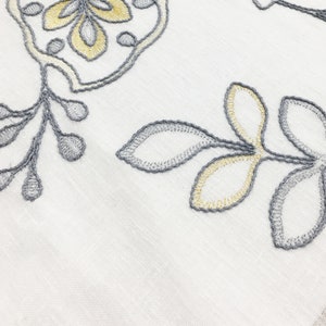 May include: White fabric with embroidered floral designs in gray and yellow. The design features leaves and flowers.