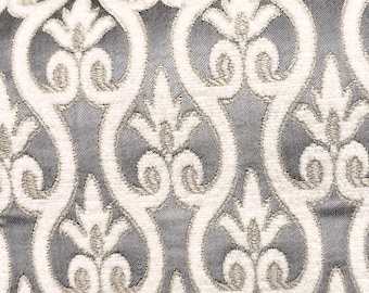 May include: Close-up of a fabric with a repeating pattern. The design features white, embroidered, scrolling floral motifs against a gray background. The texture of the fabric is visible, with the raised embroidery adding dimension to the design.