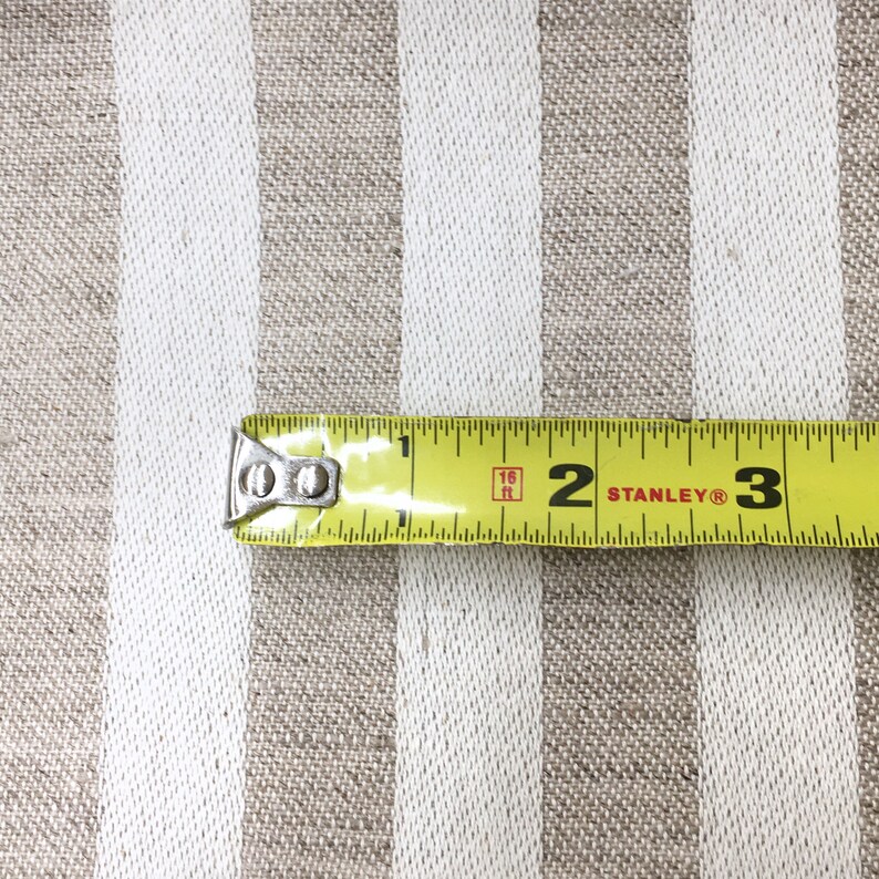 May include: A yellow measuring tape with black markings and a silver end cap. The tape reads "16 ft" and "STANLEY&reg;". The tape is measuring a beige and white striped fabric.